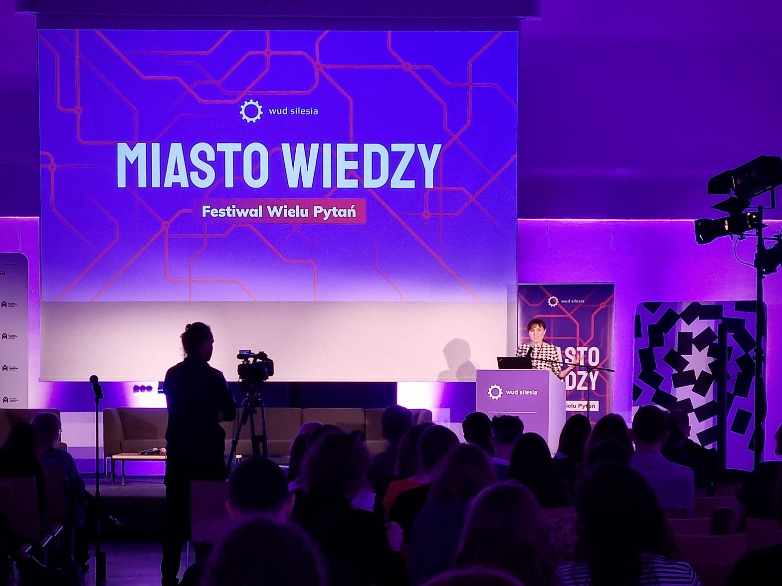 Zdjęcie sceny z konferencji. Widownia widziana od tyłu, przygaszone fioletowe światło, na mównicy kobieta. Na ekranie dużym wyświetlony slajd Miasto Wiedzy.